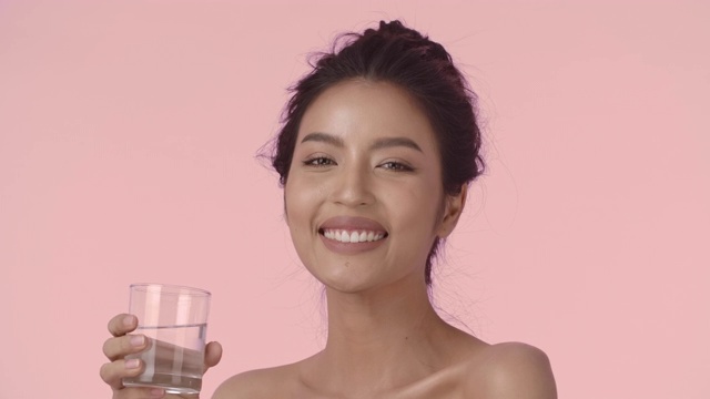 迷人的亚洲女人喝水。特写美丽的年轻亚洲女人与完美的光滑健康的皮肤喝水从一个杯子和微笑看着相机在粉红色的背景。视频素材
