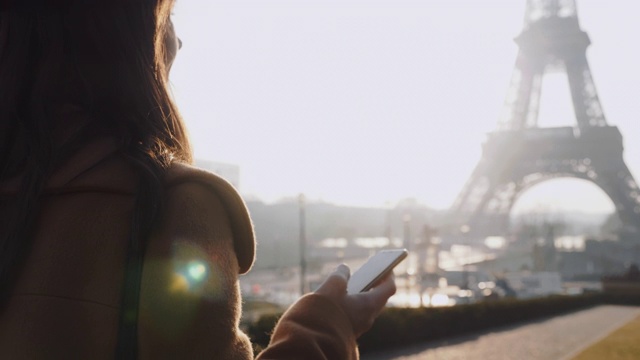 年轻美丽的女商人使用手机短信应用程序走在背光阳光下的巴黎埃菲尔铁塔看慢镜头。视频素材