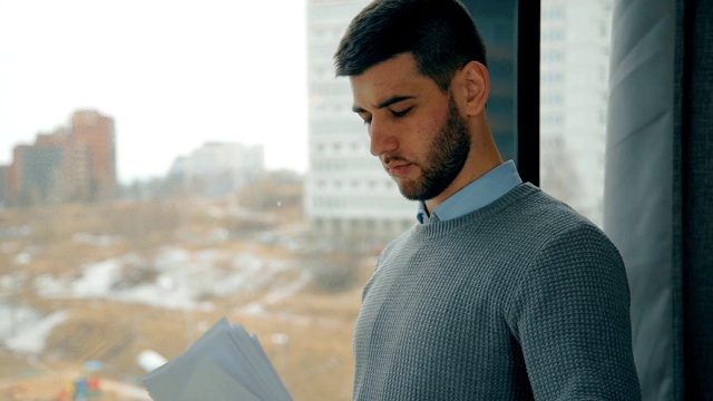 在现代化的办公室里，一个年轻人站在窗边看文件视频素材