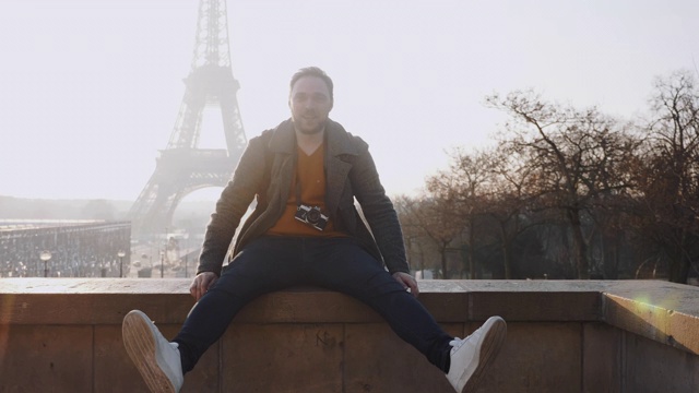 快乐的男性欧洲自由摄影师男人坐在栅栏上，微笑着在巴黎埃菲尔铁塔的风景慢镜头。视频素材