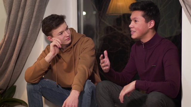 两个兴奋的男孩坐在窗台上聊天。年轻的亚洲男孩与白人朋友在室内交谈。两个大学生在家休息。视频素材