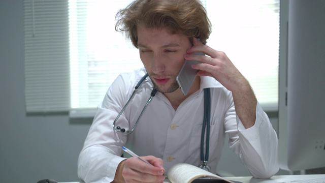 医生坐在医院柜子的桌子旁，打电话和写东西视频素材