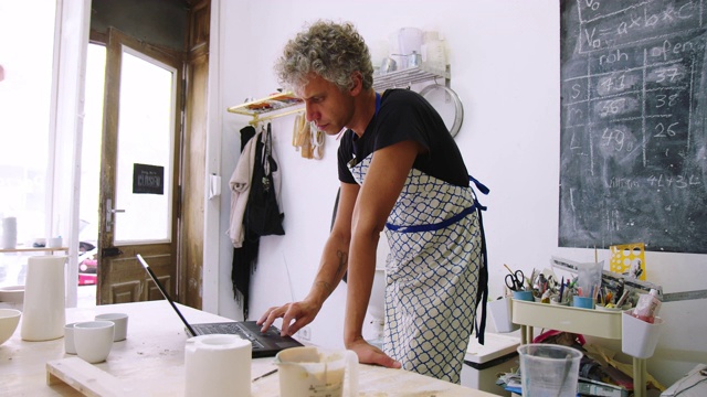 成熟的男子陶艺艺术家在艺术工作室使用笔记本电脑视频下载