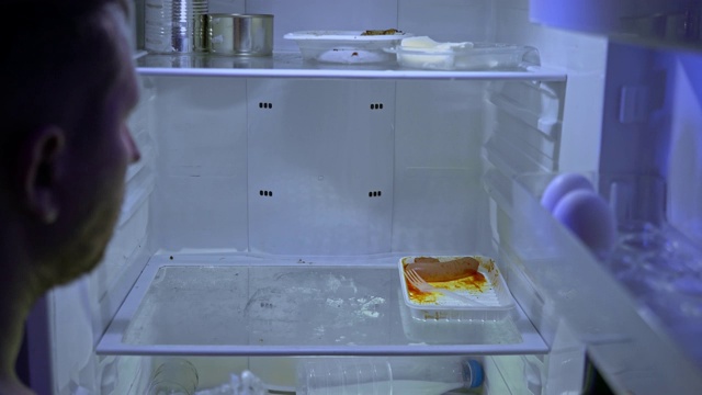 在冰箱里找食物的人。一个没刮胡子的男人在他的冰箱里挖洞，试着吃剩菜，然后关上它。视频下载
