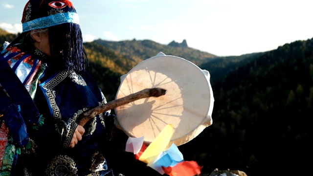 一个穿着鲜艳民族服装的老萨满在山顶敲着小手鼓进入恍惚状态。视频下载