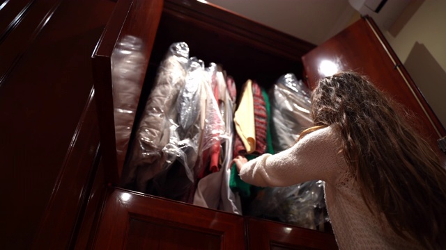深色头发的女孩在她的木制衣柜里翻找，女人在衣架上挑选衣服，决定穿什么。家居衣柜，选择问题概念视频下载
