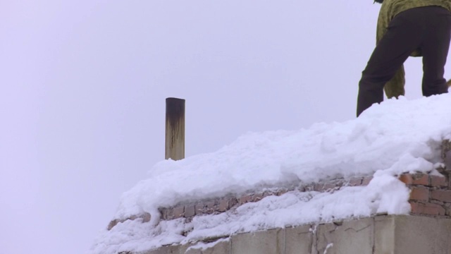 一名男性工人正在用铲子清理建筑物屋顶上的积雪。头上有落雪和冰柱的危险。视频素材
