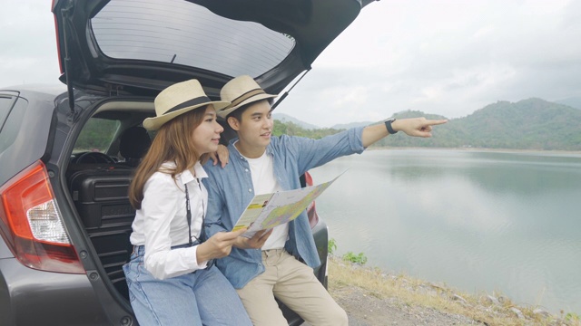 肖像的亚洲夫妇坐在汽车与男人和女人看美丽的风景和拿着地图，而度假在一起。幸福浪漫的时间。视频素材