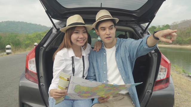 肖像的亚洲夫妇坐在汽车与男人和女人看美丽的风景和拿着地图，而度假在一起。幸福浪漫的时间。视频素材