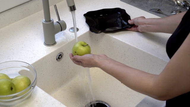 一个成年女孩在厨房水槽的水流下洗干净漂亮的青苹果，然后把它们放在一个黑色的盘子里。家庭作业。健康的饮食习惯。关闭了。4 k视频素材