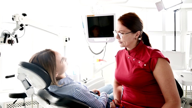 一个年轻的女孩坐在牙科诊所的椅子上，向一位戴眼镜的女牙医咨询。医生和病人的对话视频素材
