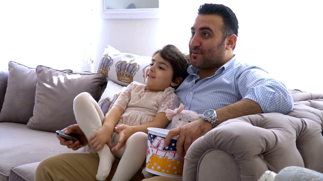 父亲和他的小女儿坐在沙发上一边看电视一边吃爆米花视频素材