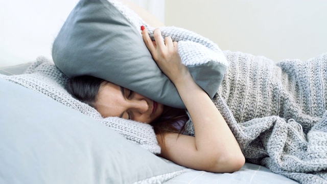 年轻急躁的女人躺在床上试图睡觉。愤怒的女孩用枕头捂住耳朵，大声的噪音打扰了她。视频素材