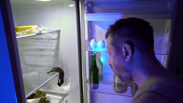 在冰箱里找食物的人。一个没刮胡子的男人在他的冰箱里挖洞，试着吃剩菜，然后关上它。视频下载