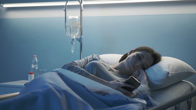 年轻的病人躺在医院的病床上，正在使用她的智能手机视频素材