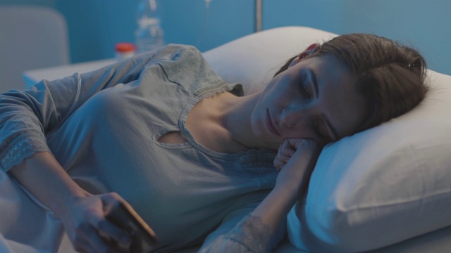 年轻的病人躺在医院的病床上，正在使用她的智能手机视频素材