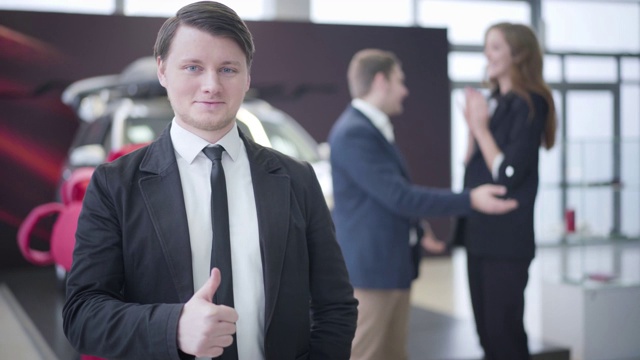 肖像自信的白人汽车经销商站在经销商和显示拇指作为年轻幸福的夫妇拥抱和旋转在背景。汽车工业，商业，贸易。视频下载