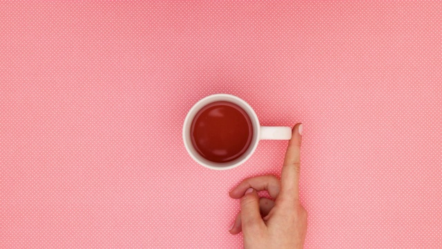 女人用手指在粉红色的背景上圈动瓷杯中的茶-停止运动视频素材
