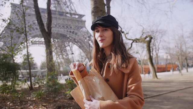 美丽快乐的欧洲女游客走在巴黎埃菲尔铁塔微笑，吃新鲜的法式长棍面包慢动作。视频素材