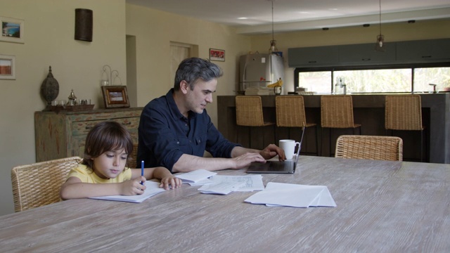慈爱的爸爸一边用笔记本电脑工作，一边喝咖啡，在儿子画画的时候对他微笑视频素材