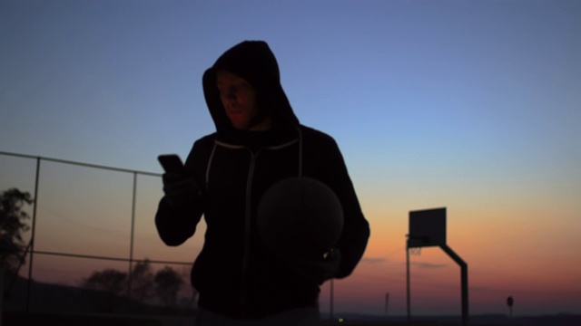 年轻男子使用智能手机，在黄昏时走出户外篮球场视频下载