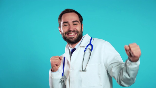 年轻英俊的医生男人在专业医疗白大褂是孤立的蓝色工作室背景。长着胡子的医生跳舞滑稽，工作成功又幸运。视频下载