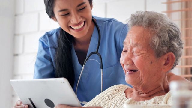 亚洲老年妇女与年轻的黑人女护士医生交谈，帮助患者在家里进行平板电脑治疗Covid-19病毒的教育。教育、医疗、护理、技术、老年护理、退休、志愿者、慈善、养老院、冠状病毒或新冠肺炎视频下载