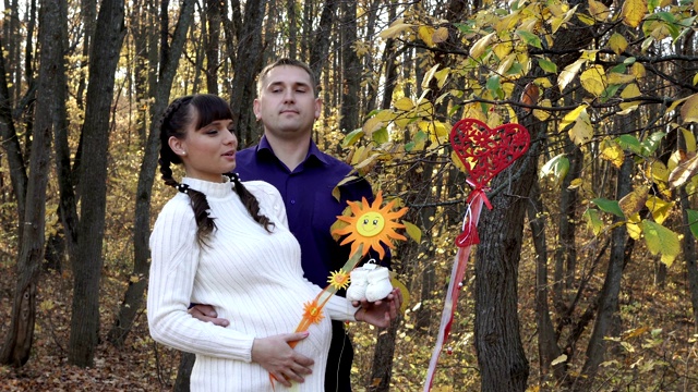 一对相爱的怀孕夫妇在秋天的森林里拥抱亲吻。一名男子和一名怀孕的小女孩摆姿势，背景是挂在树枝上的一个玩具心和一个太阳。近距离视频下载