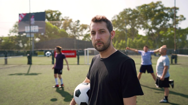 男足球运动员站在足球场上，手臂抱着球视频素材