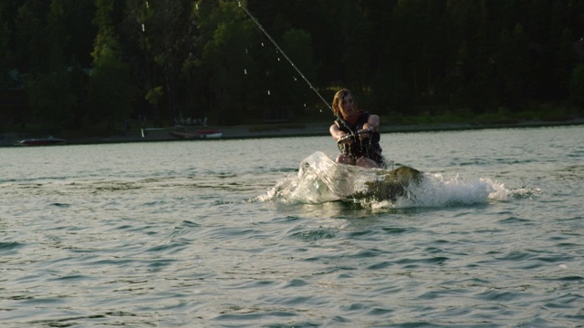 一个阳光明媚的下午，在蒙大拿维克多附近的格伦湖上，一个20多岁的白人男子站起来，跳起来，在滑水板上表演特技视频素材
