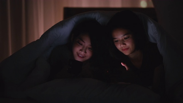 一对亚洲十几岁的女孩看手机，在晚上在卧室的毯子下一起聊天视频下载