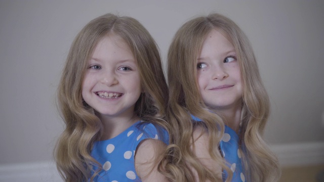 两个愉快的白人双胞胎姐妹背靠背坐着，说说笑笑。快乐的孩子们穿着相似的衣服在室内一起玩耍。生活方式,兄弟姐妹,家庭。视频素材