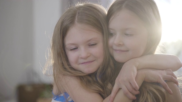 特写脸的快乐小漂亮的女孩拥抱双胞胎妹妹和亲吻她的脸颊。两个孩子在室内阳光下摆姿势。幸福、休闲、生活方式、团结。视频素材