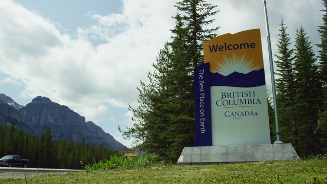 慢镜头拍摄汽车和车辆通过一个“欢迎——加拿大不列颠哥伦比亚省，地球上最好的地方”的标志旁边的加拿大落基山脉和森林在一个部分阴天视频下载
