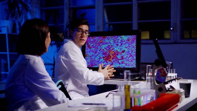 在生物化学实验室里，一名女科学家做着笔记，把装有红色液体的试管递给一名年轻的男同事，让他在电脑屏幕上看放大的微生物视频下载