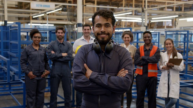 开朗的男蓝领工人戴着保护耳套在脖子上对着镜头微笑，一群员工站在后面视频下载