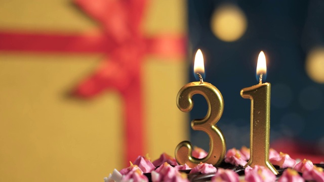 31号生日蛋糕用金色蜡烛点燃，蓝色背景礼物用黄色礼盒用红色丝带系好。特写和慢动作视频下载