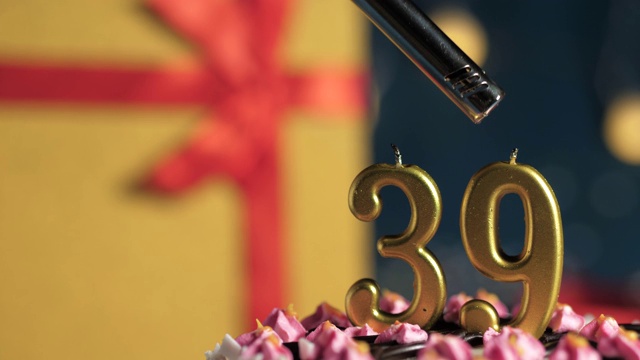 39号生日蛋糕用金色蜡烛点燃，蓝色背景礼物用黄色礼盒系上红色丝带。特写和慢动作视频下载
