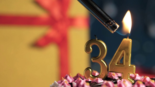 34号生日蛋糕用金色蜡烛点燃，蓝色背景的礼物用红色丝带绑在黄色盒子里。特写和慢动作视频下载