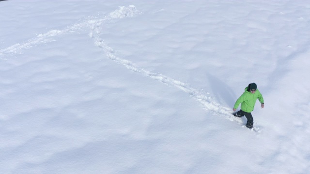冬天，一个穿着绿色羽绒服的男人走在白色的雪堆上。从无人机鸟瞰图上看到一个人坠落在雪堆上。顶部鸟瞰图人类脚印在白色蓬松的雪视频下载
