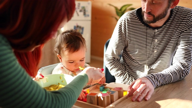 细心的父母用玩具逗他们的宝贝儿子玩，同时用花椰菜泥喂他视频素材