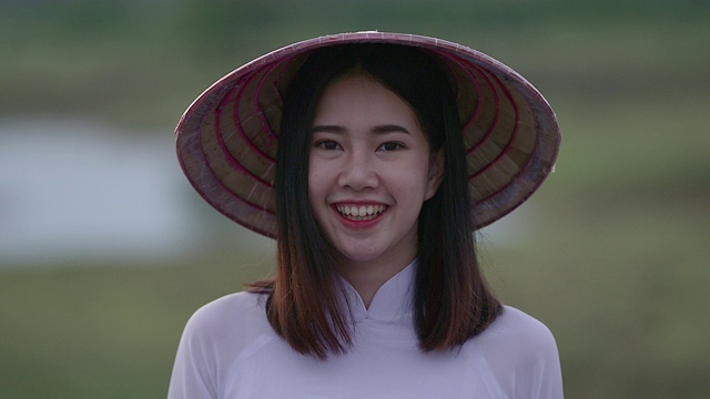 一位美丽的年轻女子穿着一件白色的连衣裙，拿着一顶竹编织的帽子。站着对着镜头笑，心情很好视频下载