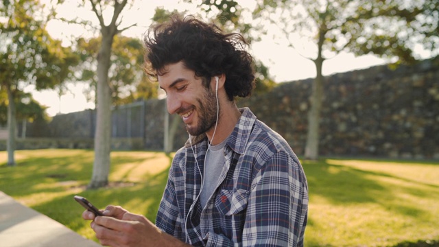 在公园里玩滑板的年轻男性戴着耳机用智能手机发短信视频素材