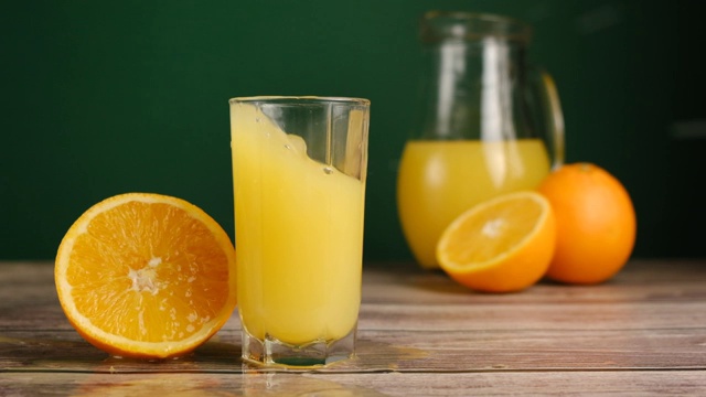 冰块落在装有橘子汁或柠檬汁的玻璃杯里，放在放着橘子的木桌上。用天然原料准备健康的早餐。慢动作视频下载