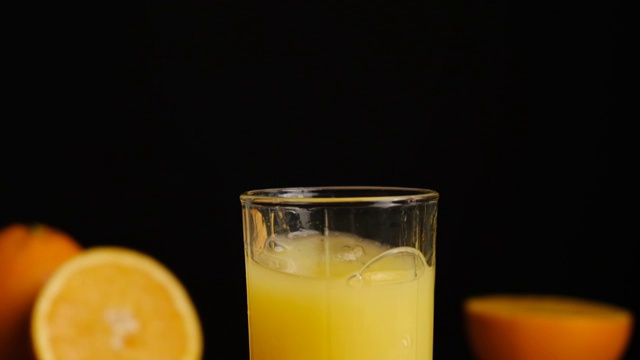 冰块落在橙汁或柠檬汁的杯子里，黑色的背景是橘子。用天然原料准备健康的早餐。缓慢的运动。视频素材