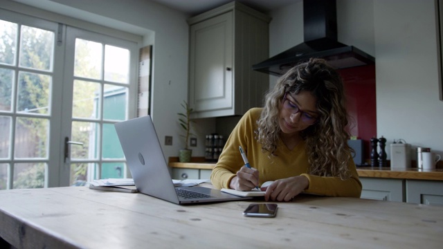 关注拉丁美洲的女性在家里使用笔记本电脑和记事本工作视频下载