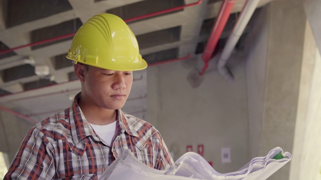 亚洲建筑工程师戴安全帽在施工现场查看图纸，工程师在现场检查建筑施工项目视频素材