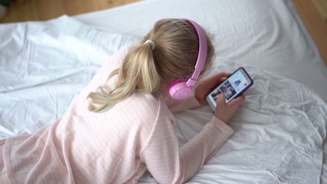 z一代的少女穿着睡衣戴着耳机躺在床上用智能手机听音乐。视频素材