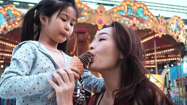 享受游乐园乐趣的概念。亚洲母亲和女儿在游乐场吃冰淇淋和一个旋转木马的背景。视频下载
