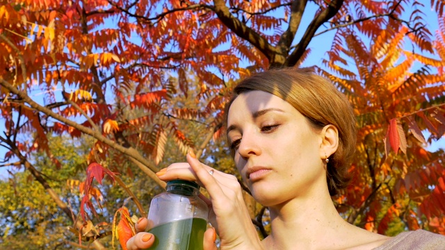 美丽的女孩在公园外面的树上喝着绿色的饮料，螺旋藻、小球藻和麦草冰沙，树的背景是红色和橙色的叶子。健康的生活方式,排毒。视频素材